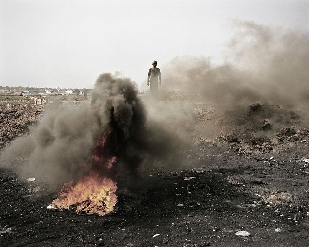 Agbobloshie, Accra, Ghana. A guy is standing in the midst of smoke, fire and residual parts of electronic equipment as he burn it to extract some copper contained inside he will resell later to gain his daily food.